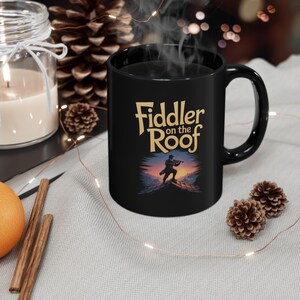 May include: Black ceramic mug featuring the text "Fiddler on the Roof" in gold lettering. The mug has a graphic of a person playing a violin on a rooftop at sunset. A candle and pinecone are in the background.