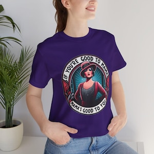 Puede incluir: Camiseta morada con un gráfico de una mujer con un vestido rojo y un sombrero. El gráfico dice "Si eres bueno con mamá, mamá es buena contigo."