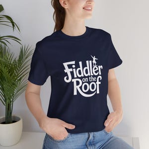 May include: Navy blue t-shirt with the words "Fiddler on the Roof" in white, along with a silhouette of a person playing a violin. The shirt is made of a soft material and has a classic crew neck.