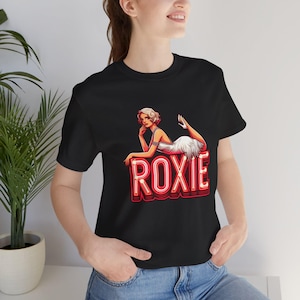 Puede incluir: Camiseta negra con un letrero de neón rojo que dice "ROXIE" y una mujer con un vestido blanco acostada encima del letrero.