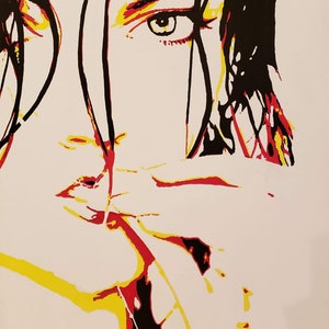 Op de afbeelding: Een close-up van een schilderij van een gezicht. De persoon heeft lang zwart haar en kijkt naar de zijkant. Het schilderij is gemaakt in popart-stijl met felle kleuren, waaronder rood, geel en zwart.
