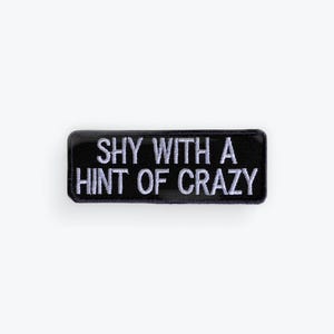 May include: A black rectangular patch with white embroidered text that reads "SHY WITH A HINT OF CRAZY". The patch has a shiny, slightly raised border.