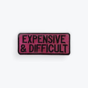 May include: A rectangular, magenta patch with black embroidered text that reads "EXPENSIVE & DIFFICULT". The patch has a black border and is likely designed to be sewn or ironed onto clothing or accessories.