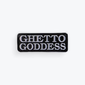 Peut inclure: Un écusson rectangulaire noir avec les mots "GHETTO GODDESS" brodés en fil argenté. L'écusson a une bordure noire et est centré sur un fond blanc.