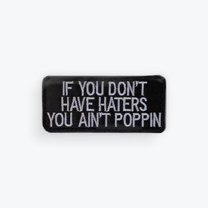 Puede incluir: Parche rectangular negro con texto bordado en blanco: "IF YOU DON'T HAVE HATERS YOU AIN'T POPPIN". El parche tiene un borde negro y está sobre un fondo blanco.