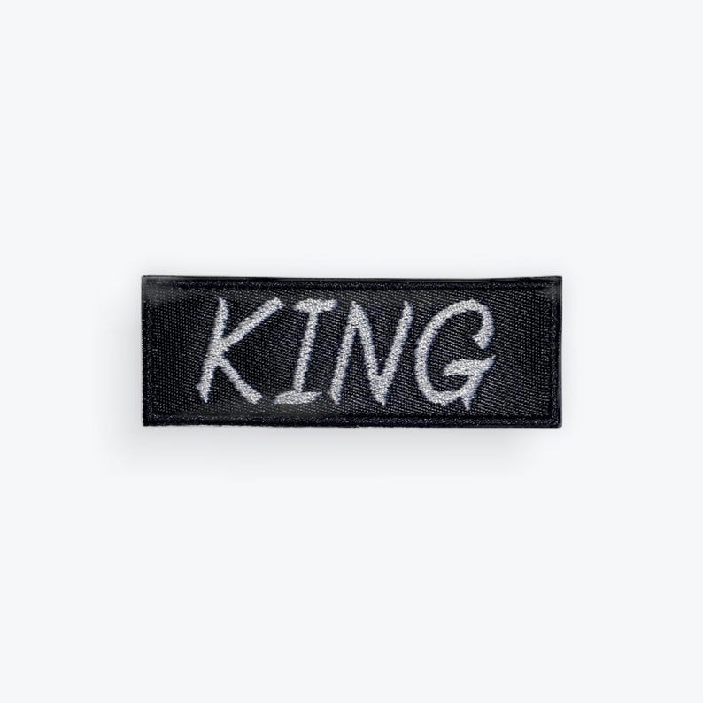 May include: A black rectangular patch with the word "KING" embroidered in silver thread. The patch has a textured appearance and is likely made of fabric. The lettering is in a handwritten style.