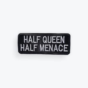 May include: A black rectangular patch with white embroidered text. The text reads "HALF QUEEN HALF MENACE". The patch has a stitched border and is likely designed to be sewn onto clothing or accessories.