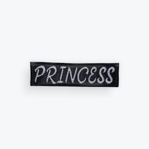 May include: A black rectangular patch featuring the word "PRINCESS" embroidered in silver thread. The patch is a simple, bold design, suitable for clothing or accessories.