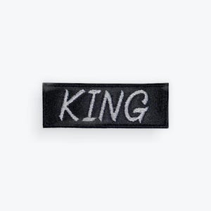 May include: A black rectangular patch with the word "KING" embroidered in silver thread. The patch has a textured appearance and is likely made of fabric. The lettering is in a handwritten style.