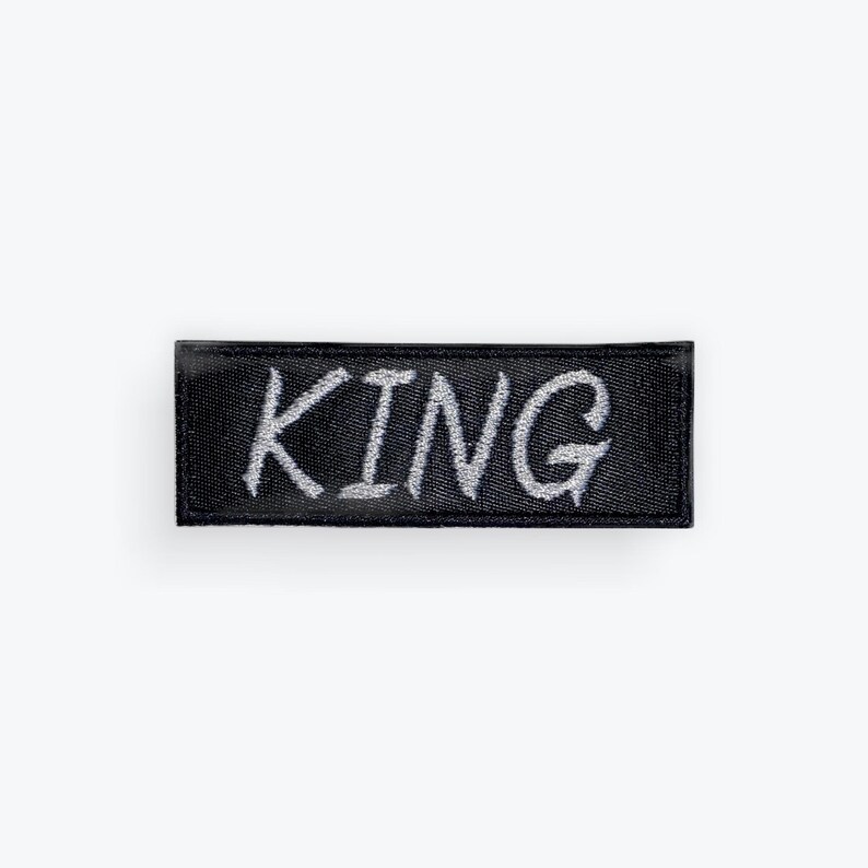 May include: A black rectangular patch with the word "KING" embroidered in silver thread. The patch has a textured appearance and is likely made of fabric. The lettering is in a handwritten style.