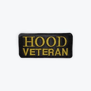 May include: A rectangular black patch with gold embroidered text that reads "HOOD VETERAN". The patch has a textured appearance and is likely made of fabric. The lettering is bold and easy to read.