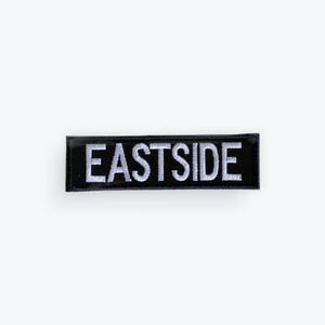 May include: A rectangular black patch with the word "EASTSIDE" embroidered in white, centered on the patch. The patch is likely made of fabric and is designed to be sewn or ironed onto clothing or accessories.