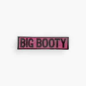May include: A rectangular, pink patch with the words "BIG BOOTY" in black, block letters. The patch has a black border and is set against a white background.