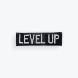 May include: A rectangular black embroidered patch featuring the white text LEVEL UP in a bold, sans-serif font. The patch has a clean, stitched border and a flat, horizontal orientation, designed for application onto fabric surfaces.