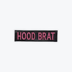 May include: Black rectangular patch with the embroidered text "HOOD BRAT" in pink. The patch is likely made of fabric and designed to be sewn or ironed onto clothing or accessories.