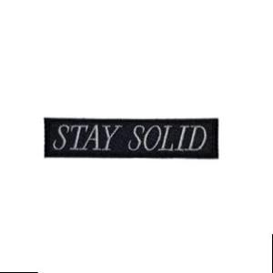 May include: A black rectangular patch with the words "STAY SOLID" embroidered in white, centered horizontally. The patch is likely made of fabric and designed to be sewn or ironed onto clothing or accessories.