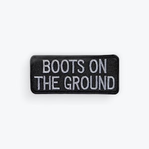 May include: A rectangular embroidered patch with a black background and a thick black border. The text BOOTS ON THE GROUND is stitched in white capital letters across the center of the patch in a bold, sans-serif font style.
