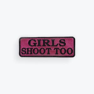 May include: A rectangular patch with a magenta background and black embroidered text. The text reads "GIRLS SHOOT TOO". The patch has a black border and is likely designed to be sewn or ironed onto clothing or accessories.