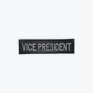 May include: A black rectangular patch with the embroidered text "VICE PRESIDENT" in white. The patch is likely made of fabric and is designed to be sewn or attached to clothing or accessories.