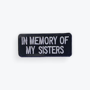Puede incluir: Un parche rectangular negro con texto bordado blanco que dice "IN MEMORY OF MY SISTERS". El parche tiene un borde brillante y está diseñado para ser cosido en ropa o accesorios. Un artículo conmemorativo.
