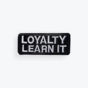 May include: Black rectangular patch with the embroidered text "LOYALTY LEARN IT" in white. The patch has a black border and is set against a white background. The text is in a bold, sans-serif font.