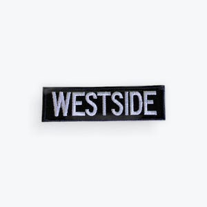 May include: Black rectangular patch with the word "WESTSIDE" embroidered in white capital letters. The patch has a glossy finish and is set against a white background. The patch is likely for clothing or accessories.