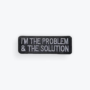 May include: Black rectangular patch with the embroidered text "I'M THE PROBLEM & THE SOLUTION" in white. The patch has a black border and is likely made of fabric.
