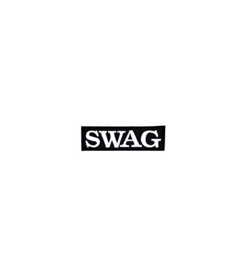 Może przedstawiać: Czarna prostokątna naszywka z napisem "SWAG" w białych, pogrubionych, wielkich literach. Naszywka jest wyśrodkowana na białym tle. Naszywka jest prawdopodobnie przeznaczona na odzież lub akcesoria.