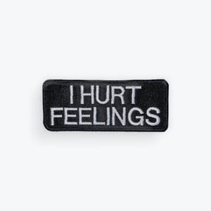 May include: A black rectangular patch with the words "I HURT FEELINGS" embroidered in white. The patch has a black border and is likely designed to be sewn or ironed onto clothing or accessories.