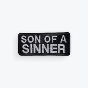 May include: A rectangular black embroidered patch with the words "SON OF A SINNER" in white thread. The patch has a black border and is likely designed to be sewn or ironed onto clothing or accessories.
