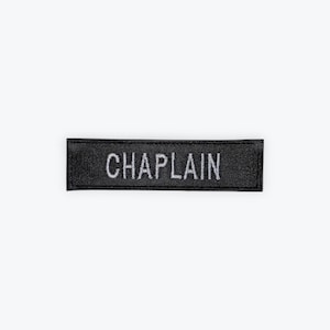 May include: Black rectangular patch with the word "CHAPLAIN" embroidered in white. The patch is centered and isolated on a white background. The text is in a sans-serif font.