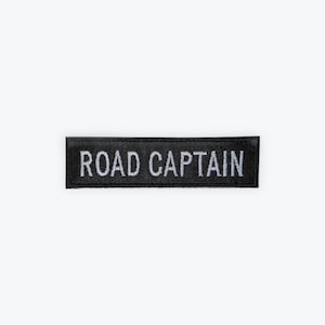 May include: A black rectangular patch with the words "ROAD CAPTAIN" embroidered in white. The patch is likely made of fabric and is designed to be sewn or ironed onto clothing or gear. The text is in a sans-serif font.