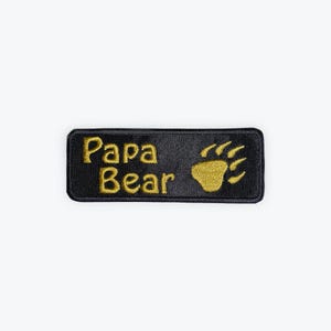 May include: Black rectangular patch with gold embroidered text and a bear paw print. The text reads "Papa Bear". The patch is likely designed to be sewn or ironed onto clothing or accessories.