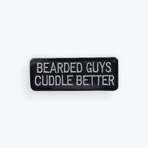 May include: A rectangular black patch with white embroidered text that reads "BEARDED GUYS CUDDLE BETTER". The patch has a black border and is likely designed to be sewn onto clothing or accessories.