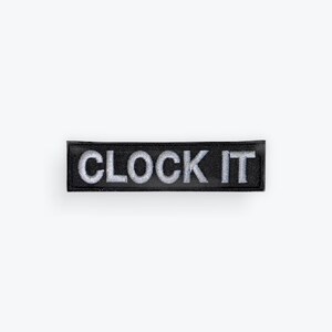 May include: A rectangular black patch with the words "CLOCK IT" embroidered in silver thread. The patch is centered on a white background, showcasing the text prominently.