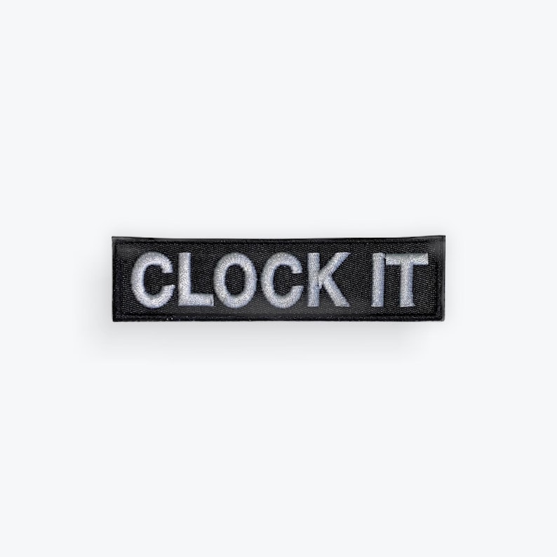 May include: A rectangular black patch with the words "CLOCK IT" embroidered in silver thread. The patch is centered on a white background, showcasing the text prominently.