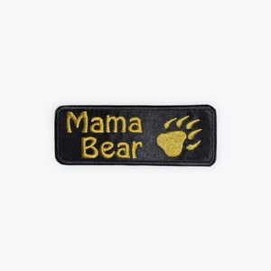 May include: Black rectangular patch with gold embroidered text and a bear paw print. The text reads "Mama Bear" in a bold, sans-serif font. The bear paw print is located to the right of the text.