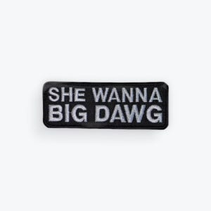 May include: A rectangular black embroidered patch with rounded corners featuring the white text SHE WANNA BIG DAWG in two lines. The patch has a thick black border and a textured fabric surface with clear, bold lettering.
