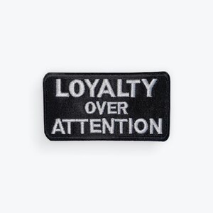 May include: Black rectangular patch with white embroidered text that reads "LOYALTY OVER ATTENTION". The patch has a black border and is likely designed to be sewn or ironed onto clothing or accessories.