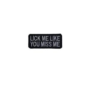 May include: Black rectangular patch with white text that reads "LICK ME LIKE YOU MISS ME". The patch is likely made of fabric and is designed to be sewn or ironed onto clothing or accessories.