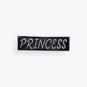 May include: A black rectangular patch with the word "PRINCESS" embroidered in silver thread. The patch is designed to be sewn or ironed onto clothing or accessories.