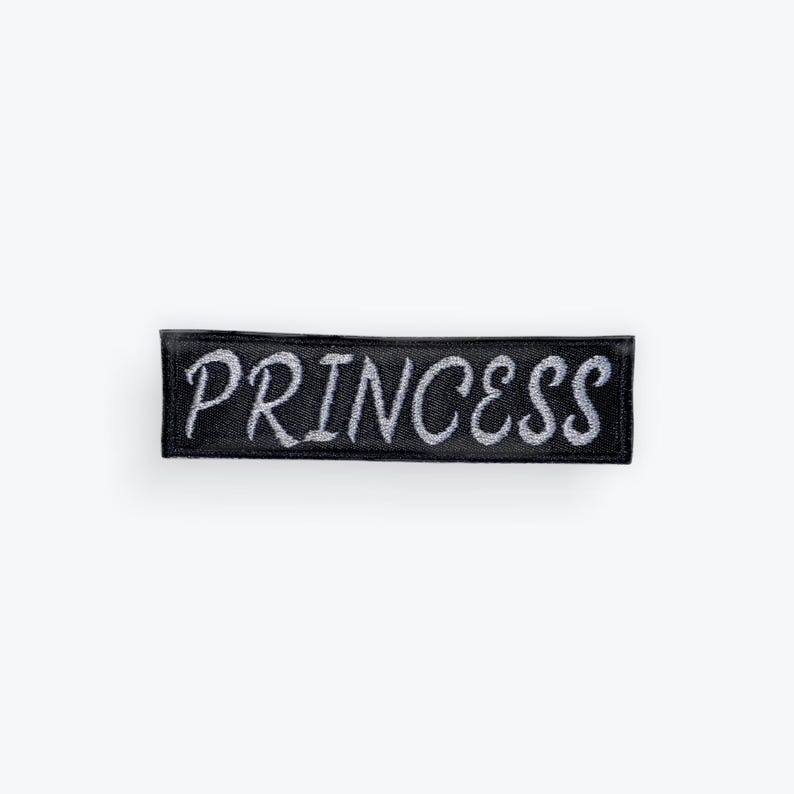 May include: A black rectangular patch with the word "PRINCESS" embroidered in silver thread. The patch is designed to be sewn or ironed onto clothing or accessories.