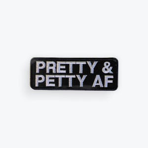 May include: A rectangular black embroidered patch with the words "PRETTY & PETTY AF" in white, stylized, capital letters. The patch has a black border and is set against a white background.