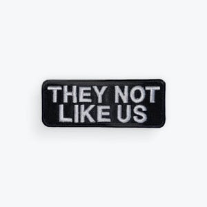 May include: Black rectangular patch with the embroidered text "THEY NOT LIKE US" in white. The patch has a black border and is set against a white background. The patch is a clothing accessory.