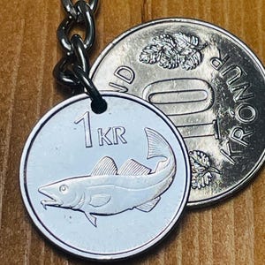 Könnte beinhalten: Ein silberfarbener, runder Schlüsselanhänger mit einem geprägten Fisch und der Aufschrift 1 KR, befestigt an einer Metallkette. Daneben liegt eine größere silberfarbene Münze mit der Aufschrift 10 KRÓNUR und einem geprägten Blattmotiv.