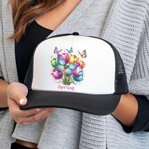 May include: A black and white trucker hat with a white front panel featuring a colorful watercolor design of tulips and butterflies. The text "spring" is printed below the design.