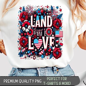 Może przedstawiać: Biała koszulka z patriotycznym wzorem. Wzór zawiera napis "LAND THAT I LOVE" otoczony czerwonymi, białymi i niebieskimi elementami kwiatowymi, gwiazdami, fajerwerkami i sercem z amerykańską flagą.