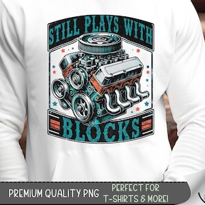 May include: A white t-shirt with a blue and red graphic of a car engine with the text "Still Plays With Blocks".