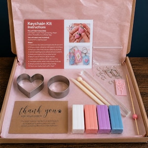 May include: A craft kit for making keychains, presented in a box. It contains clay, tools, cutters, and instructions. The kit includes a heart-shaped cutter, various coloured clay blocks, and a thank you card.
