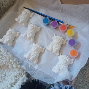 May include: A craft kit containing five white unpainted ceramic highland cow figurines, a blue paintbrush, and a strip of six small paint pots in blue, purple, orange, yellow, purple, and orange, all arranged on white tissue paper inside a box.